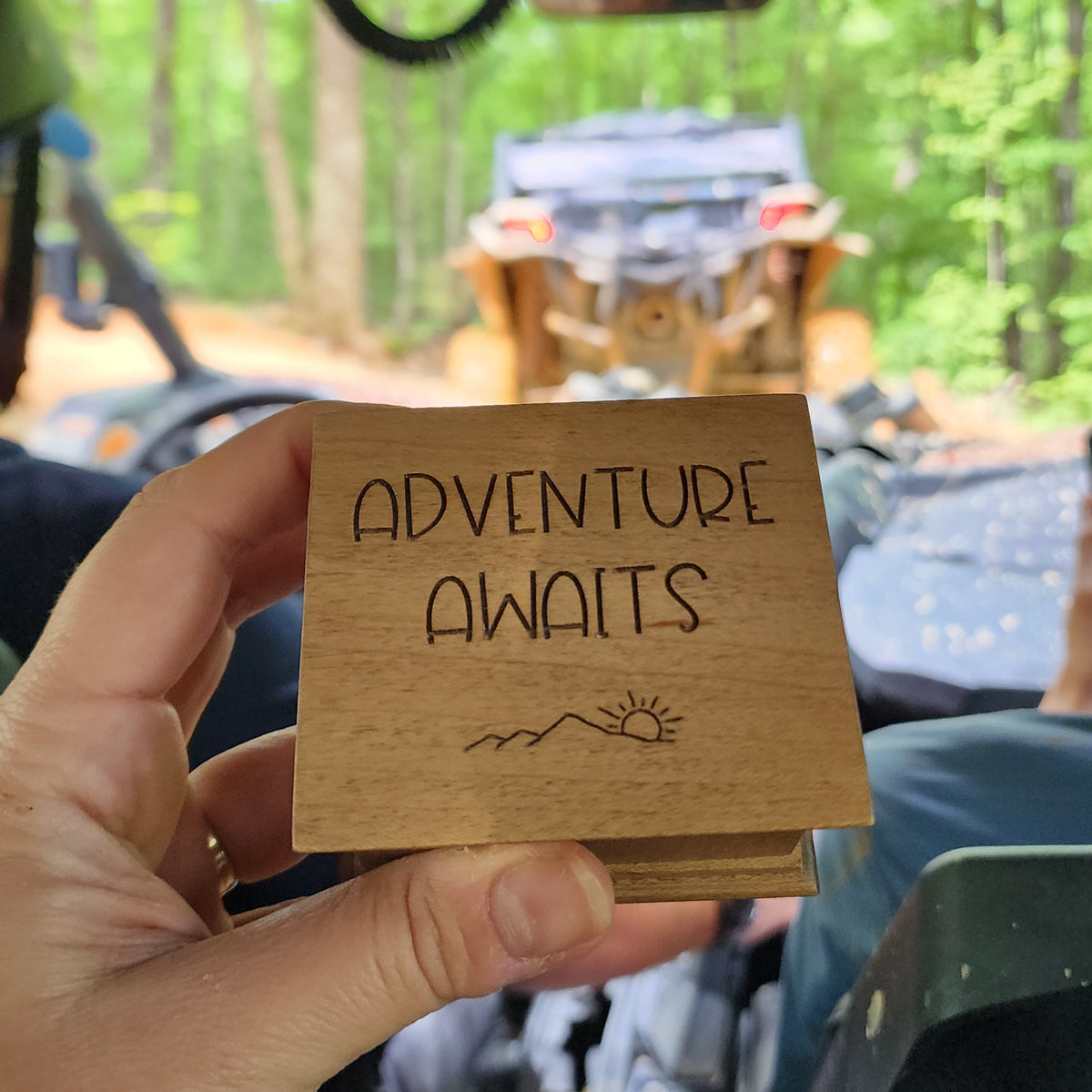Hand holding a wooden music box with 'Adventure Awaits' text inside an ATV and another ATV in the forest setting