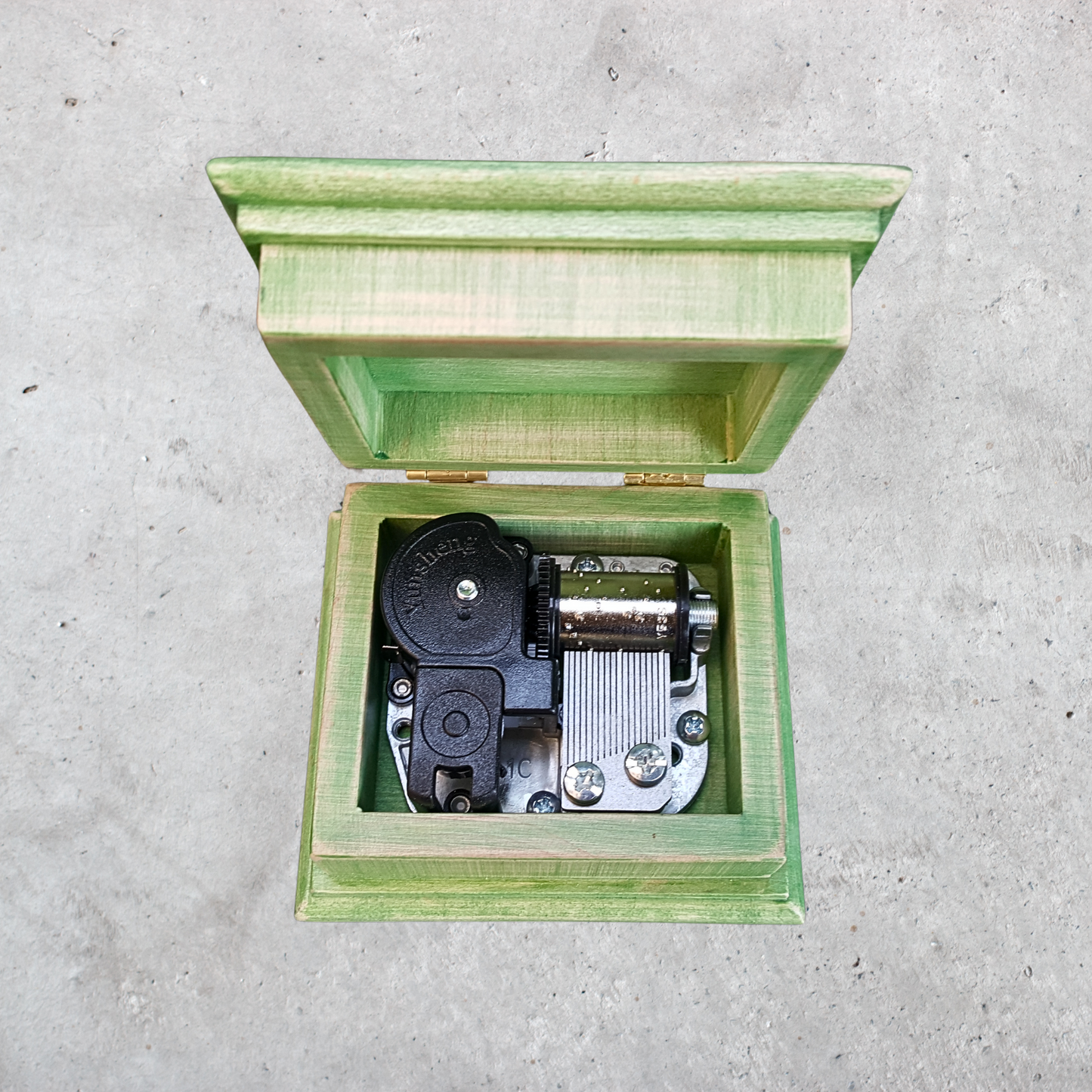 Green wooden music box with open lid on a gray concrete surface