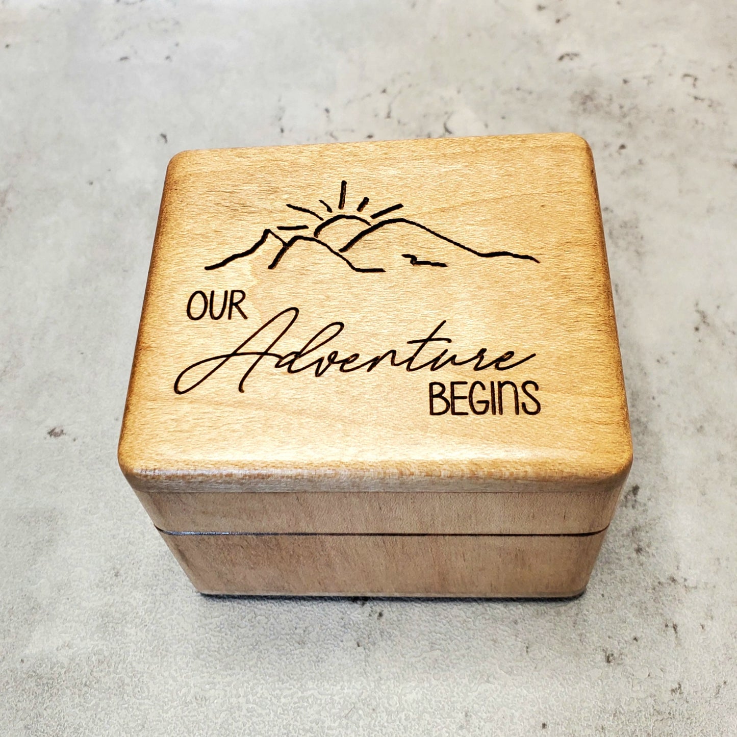 A wooden ring box with an engraved design that includes mountain silhouettes and the phrase 'Our Adventure Begins'. Plays your song in music box sound and choose from 9 different colors.