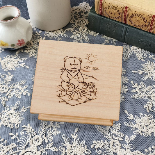 wooden music box in maple with a teddy bear sitting on a picnic blanket with mountains and sun behind him, choose song, and color, add personalized engravings