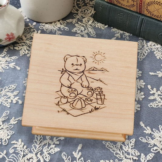 wooden music box in maple with a teddy bear sitting on a picnic blanket with mountains and sun behind him, choose song, and color, add personalized engravings