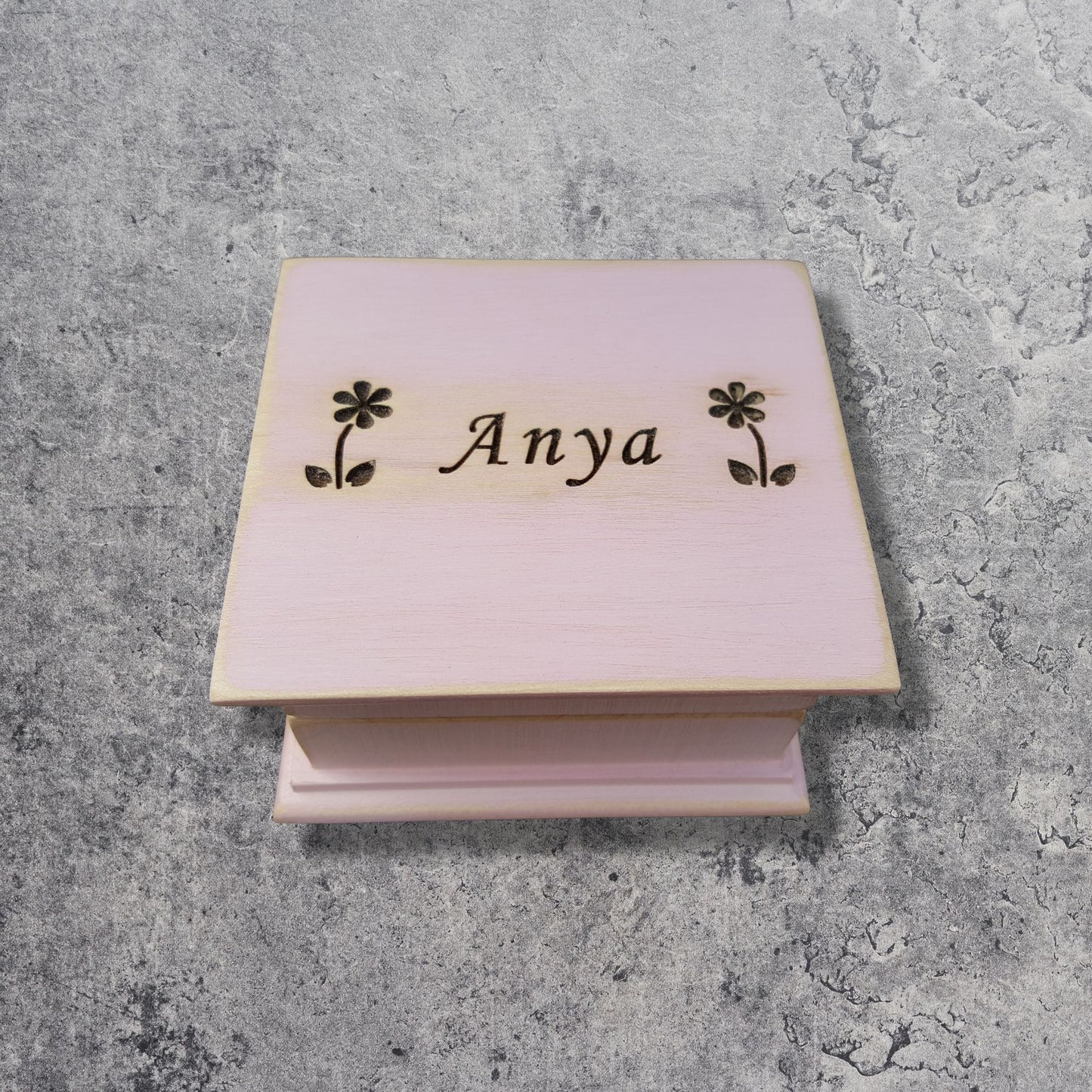 Pink wooden music box with 'Anya' and floral designs on a gray concrete surface