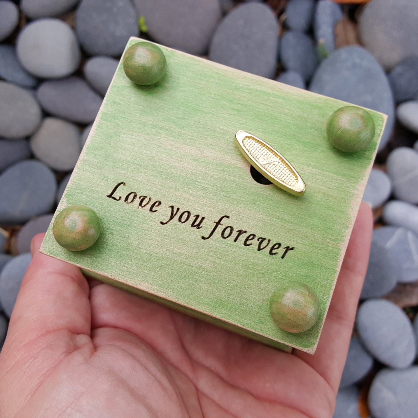 Green wooden box with 'Love you forever' engraving on the bottom side showing the 4 little button feet and the wind up key, being held over pebbles