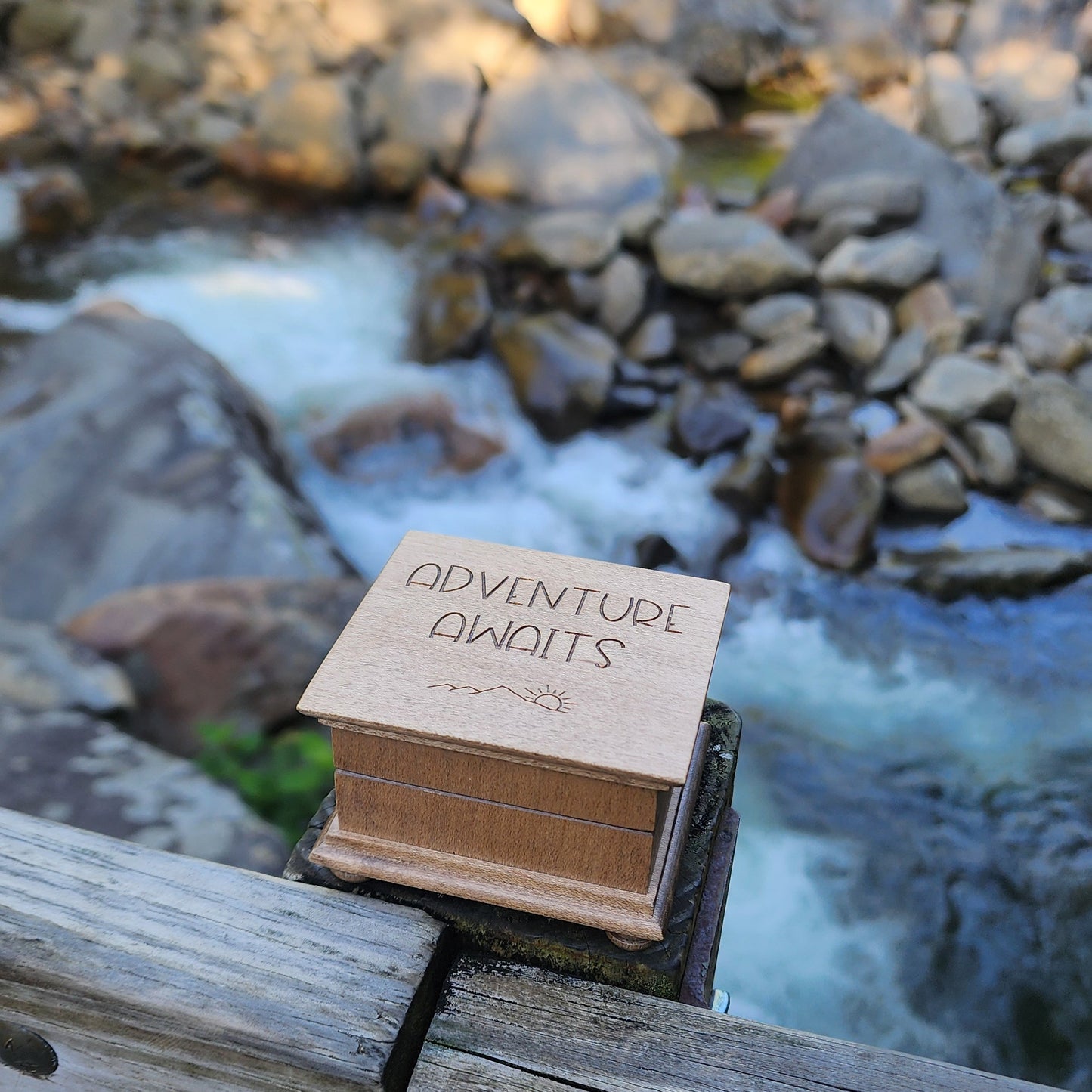 Small wooden music box labeled 'Adventure awaits' attached to a wooden post with a blurred natural background with waterwall and rocks