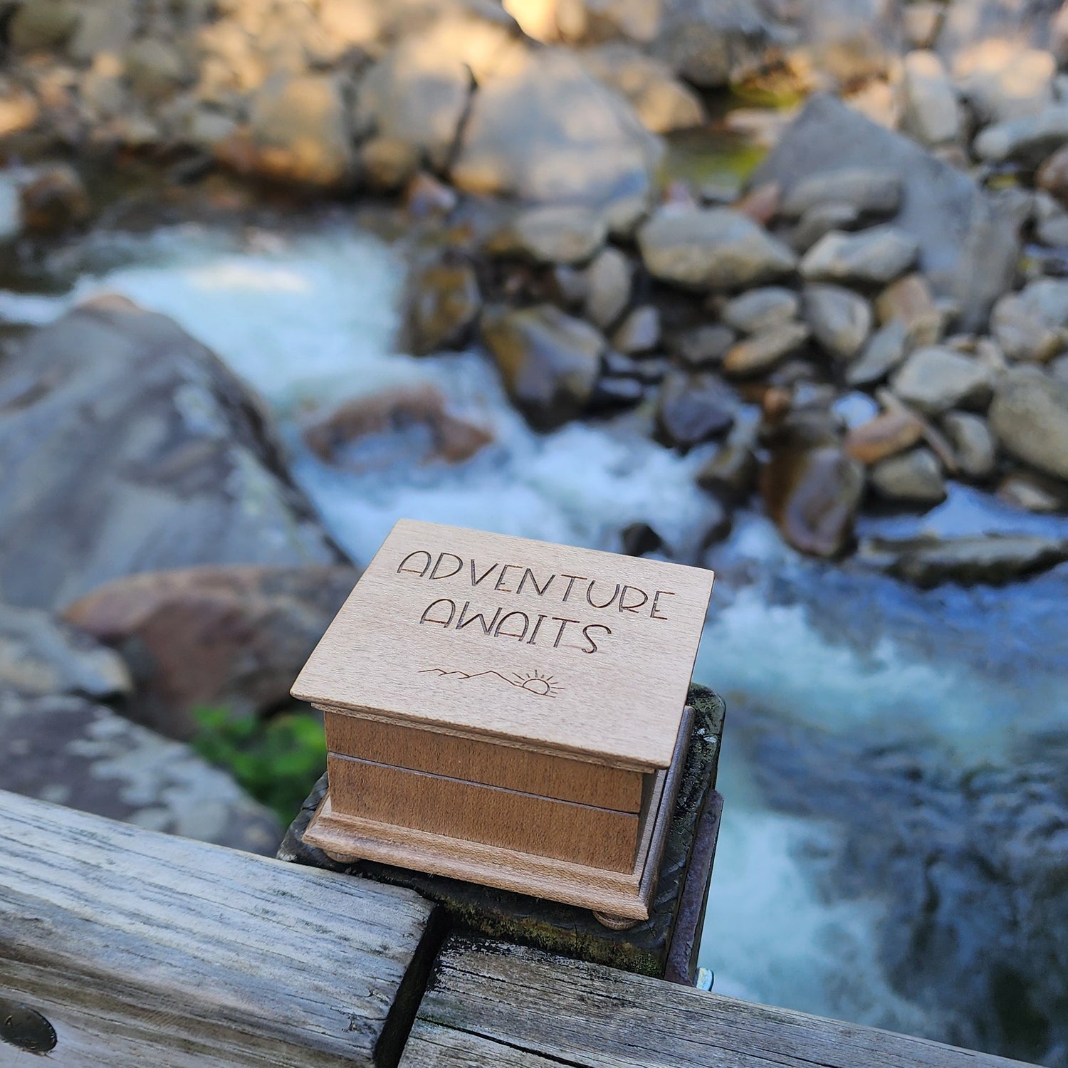 Small wooden music box labeled 'Adventure awaits' attached to a wooden post with a blurred natural background with waterwall and rocks