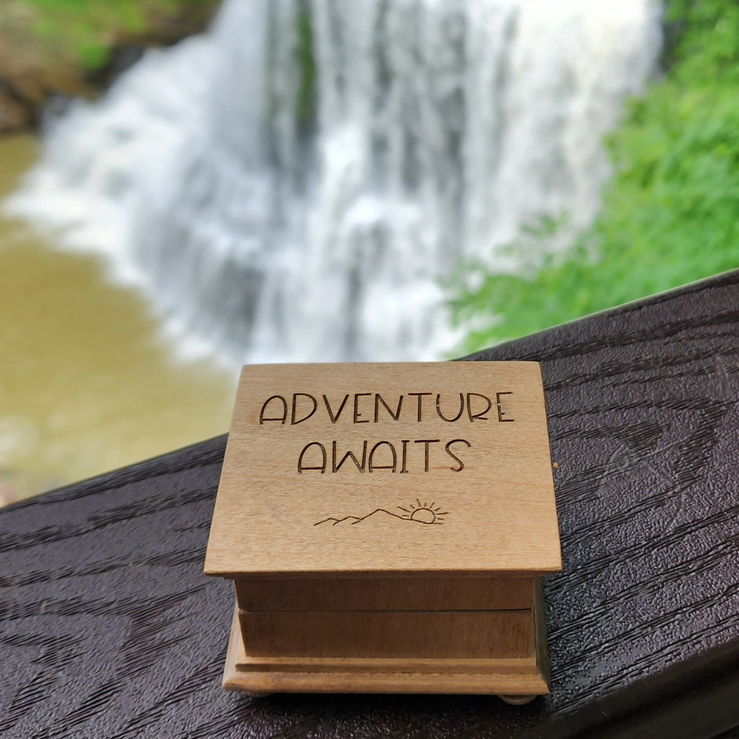 Wooden music box with 'Adventure Awaits' text in front of a waterfall