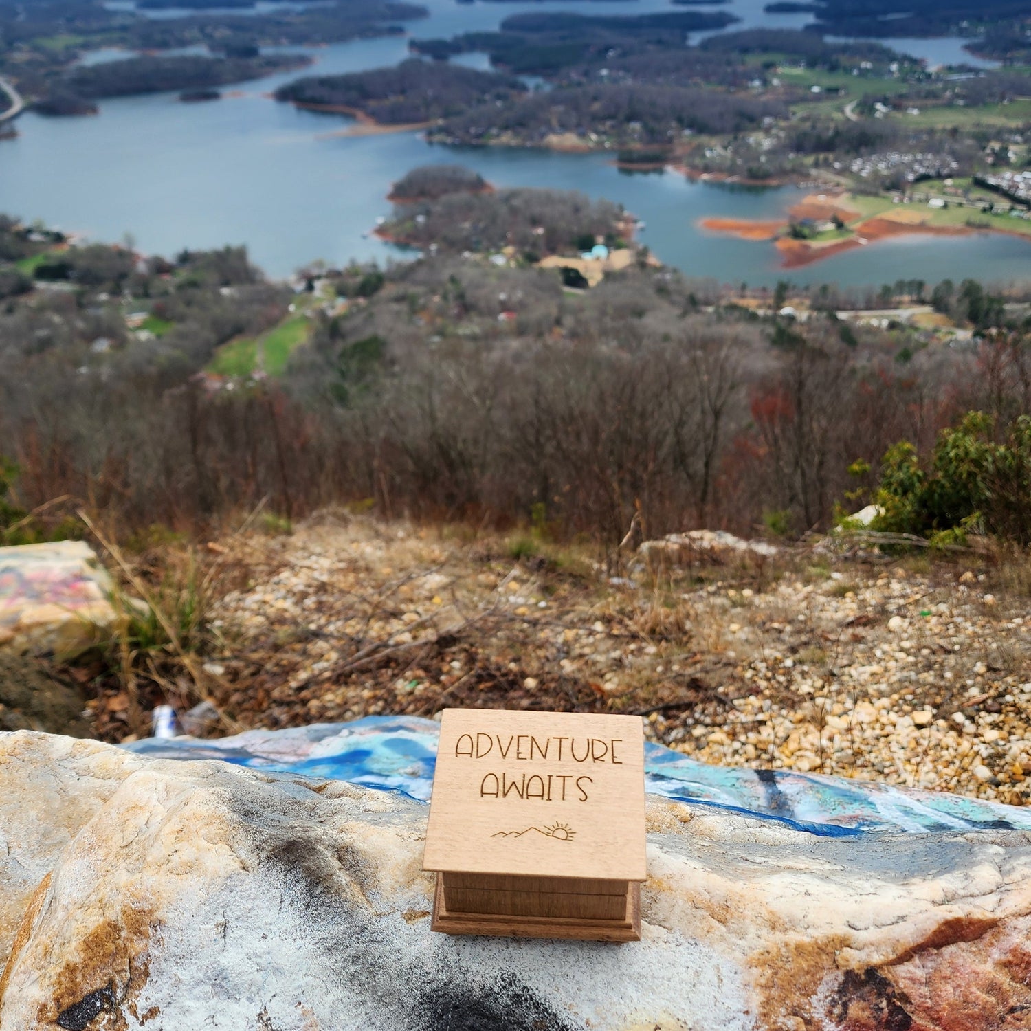 View from a high vantage point over a landscape with a music box engraved with Adventure Awaits sitting on a rock.