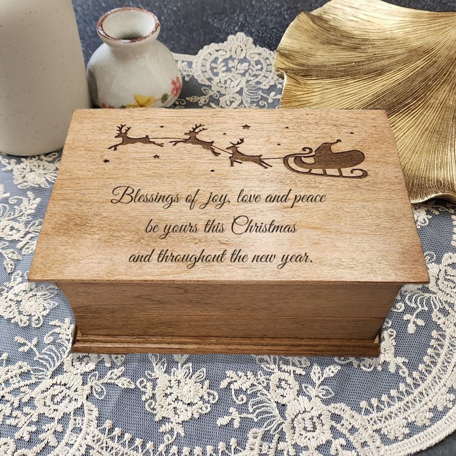 Wooden music box with Christmas message and a Santa Claus on his sleigh flying over the quote on a lace tablecloth with vases and leaves.