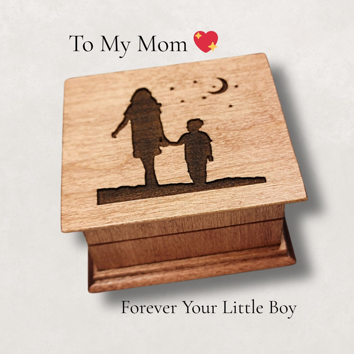 Wooden music box with engraved silhouette of a woman and son walking with moon and stars above them, text 'To My Mom' and 'Forever Your Little Boy' on a light background.