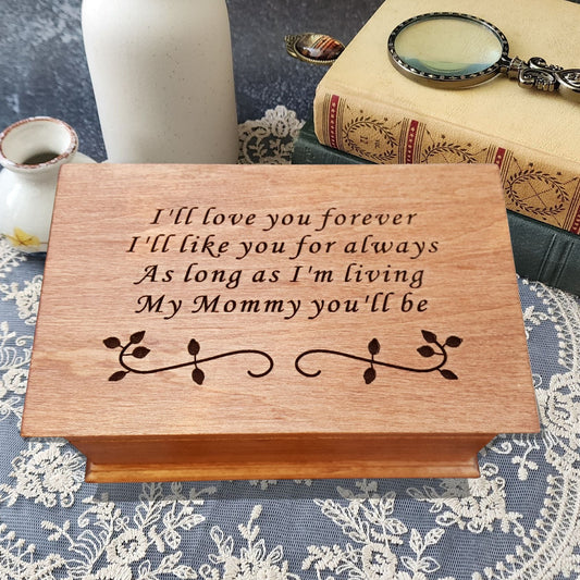 Handmade wooden music box engraved with the quote “I’ll love you forever, I’ll like you for always, as long as I’m living, my mommy you’ll be,” displayed on smooth river stones — sentimental personalized jewelry box for mom.
