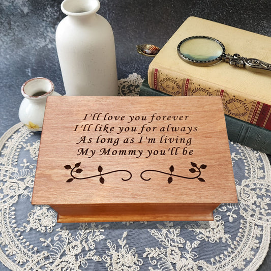 Handmade wooden music box engraved with the quote “I’ll love you forever, I’ll like you for always, as long as I’m living, my mommy you’ll be,” displayed on smooth river stones — sentimental personalized jewelry box for mom.