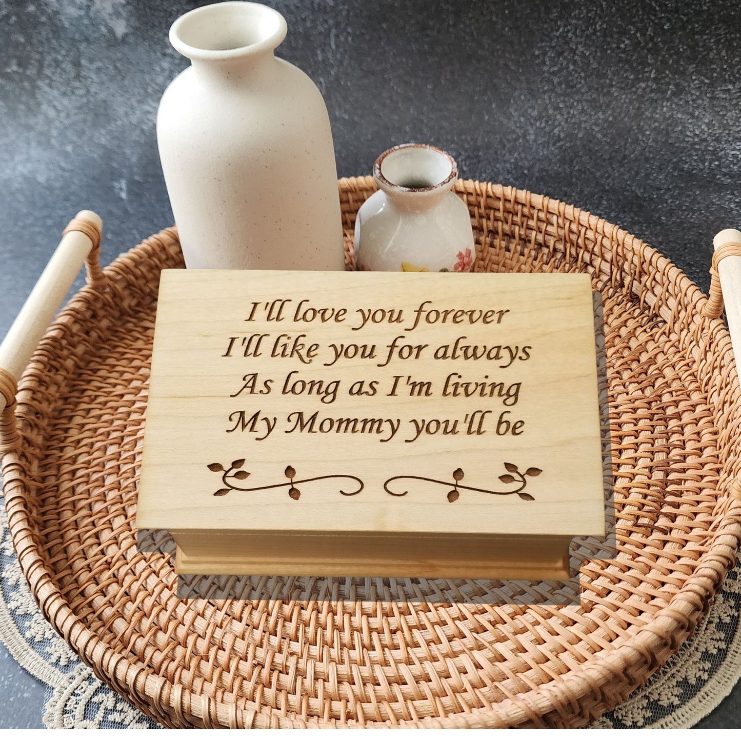 Handmade wooden music box engraved with the quote “I’ll love you forever, I’ll like you for always, as long as I’m living, my mommy you’ll be,” displayed on smooth river stones — sentimental personalized jewelry box for mom.
