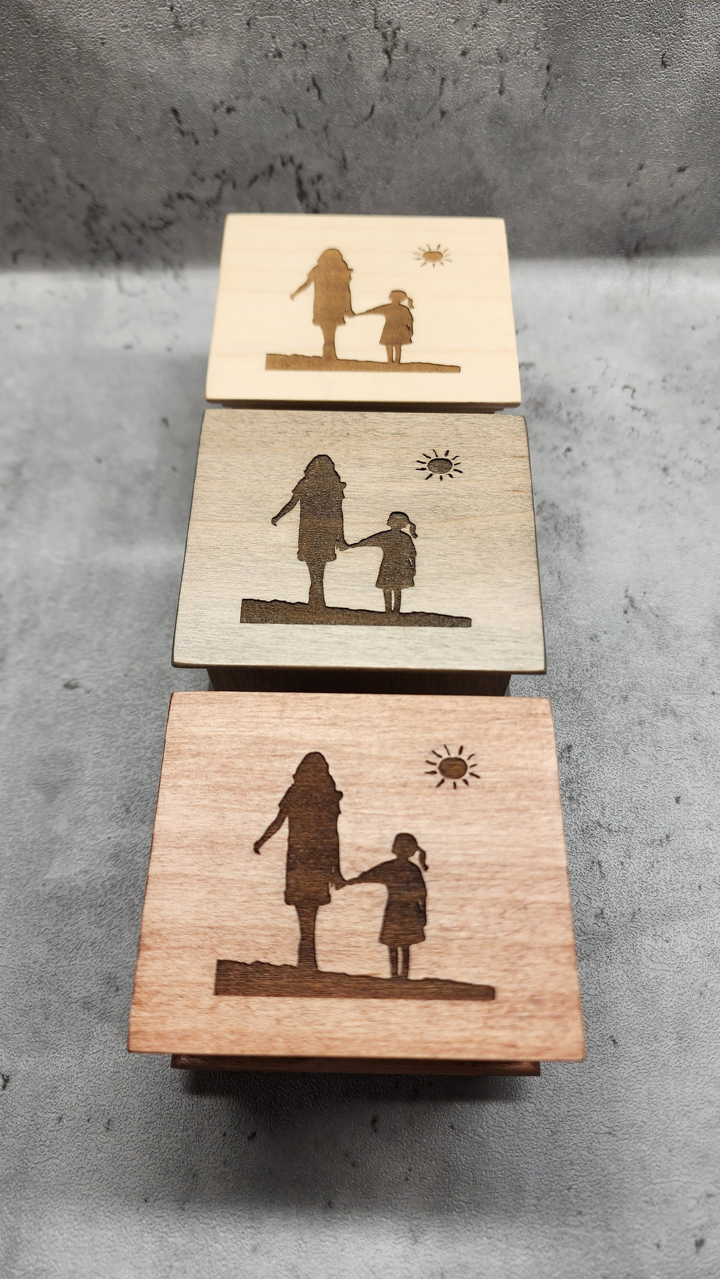 Three wooden music boxes in different colors engraved with the same silhouettes of mom and daughter holding hands and a sun shining above them on a concrete surface