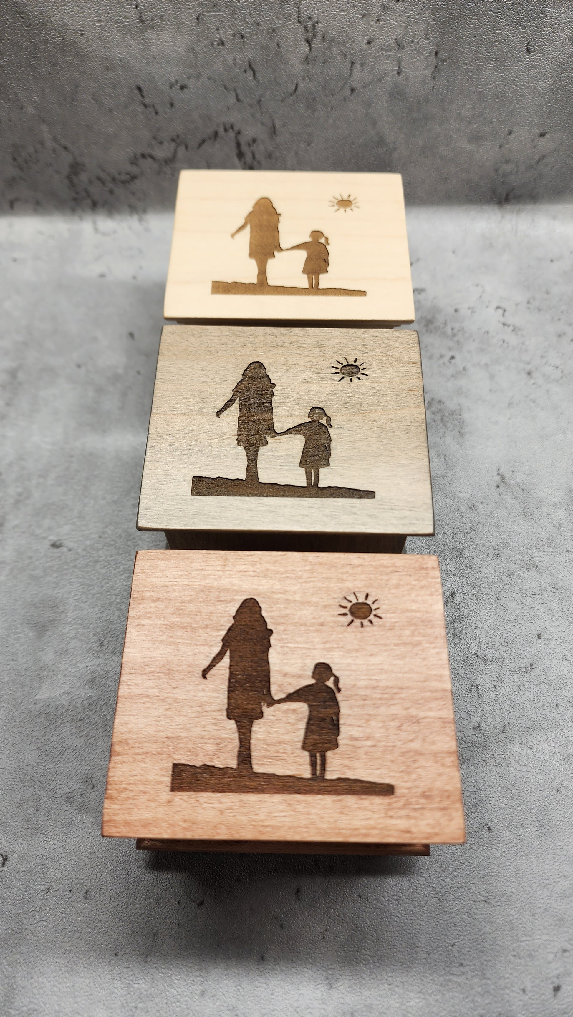Three wooden music boxes in different colors engraved with the same silhouettes of mom and daughter holding hands and a sun shining above them on a concrete surface