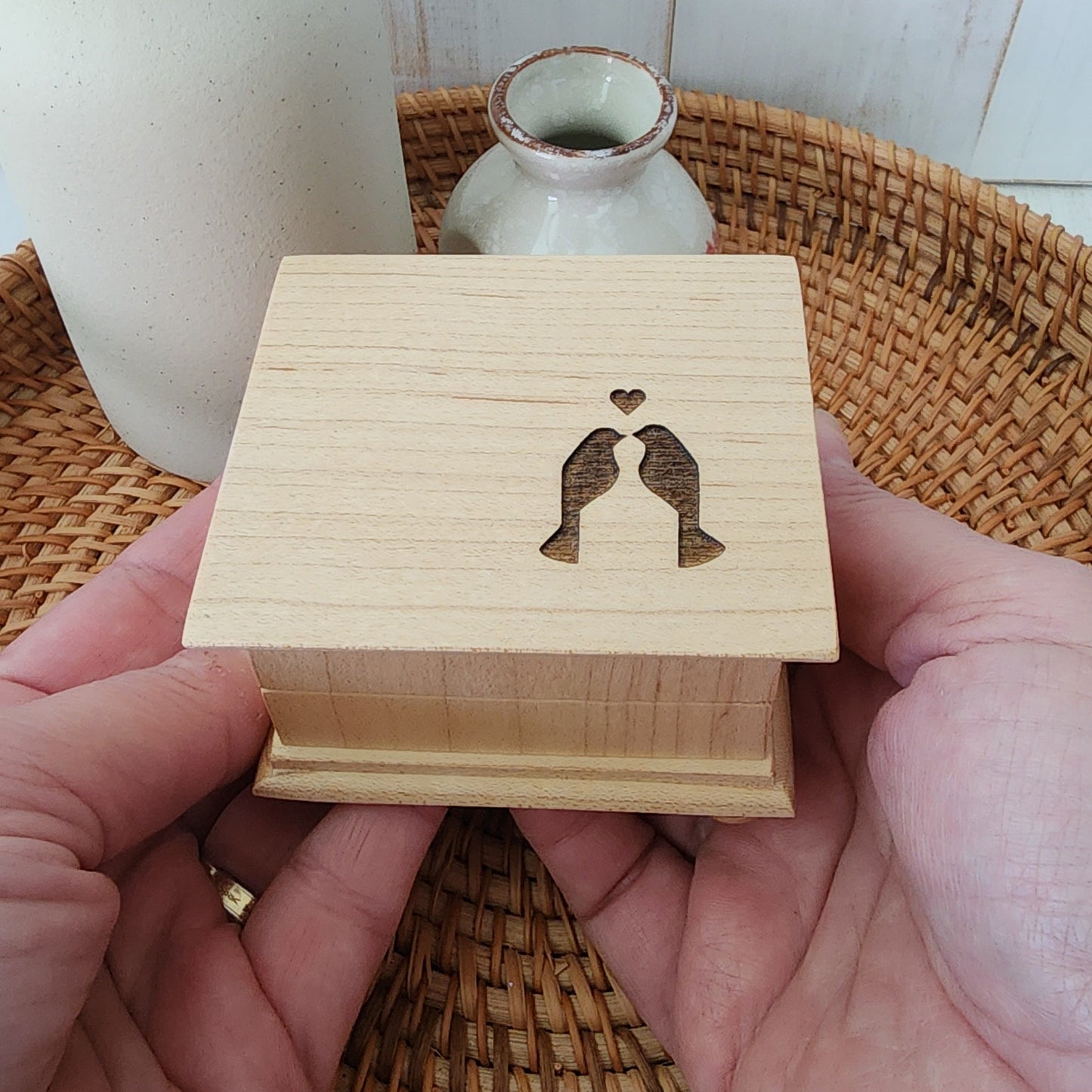 Wooden music box with laser-cut design of two birds in a heart, held in hands against a wicker basket and ceramic vase background.