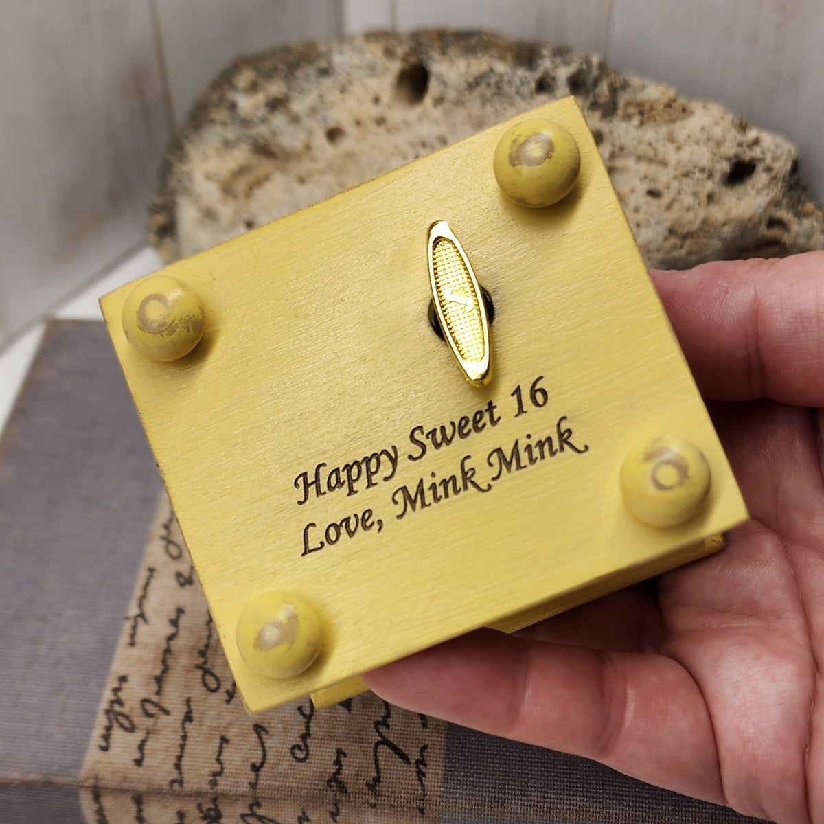 Hand holding a vintage yellow wooden music box with engraved text on a wooden surface