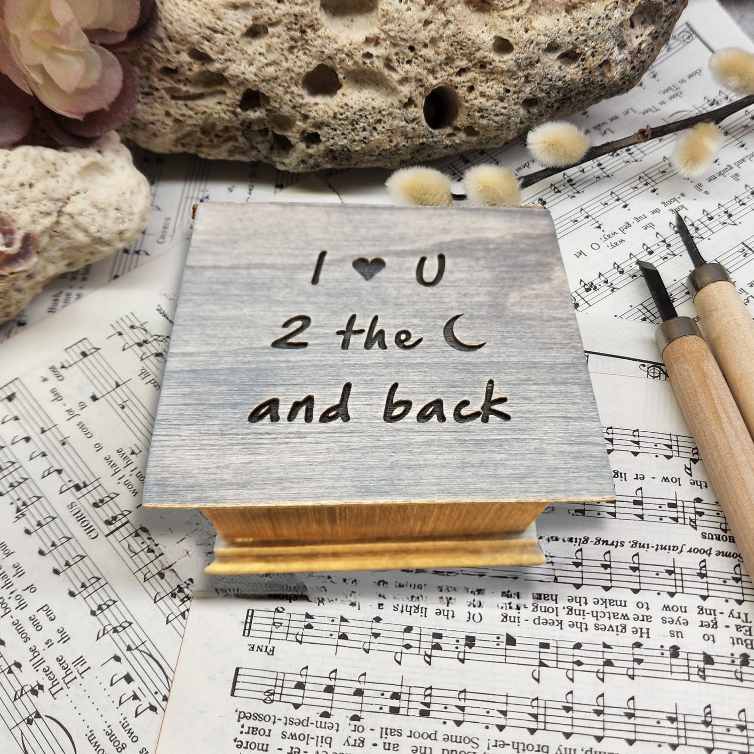 Wooden block with 'I ❤️ U 2 the moon and back' text on sheet music with a rock and flowers.
