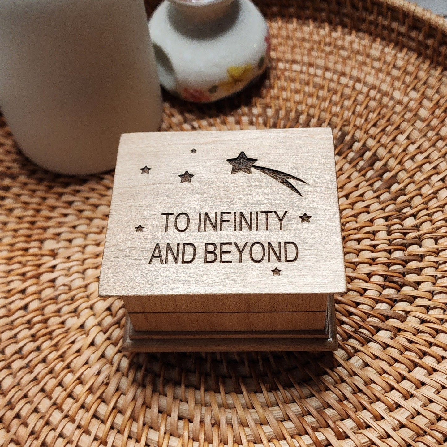 Small wooden music box with “To Infinity and Beyond” engraved on the lid, with stars and a shooting star, sitting on woven tray
