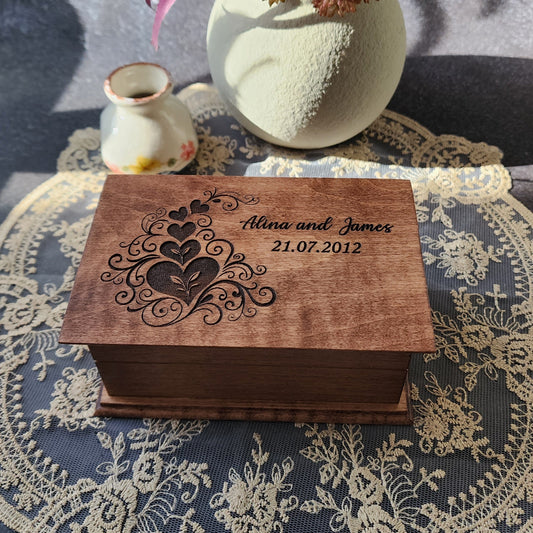 Wooden box with heart engraved design with your names and date, on a textured surface with flowers and a vase.