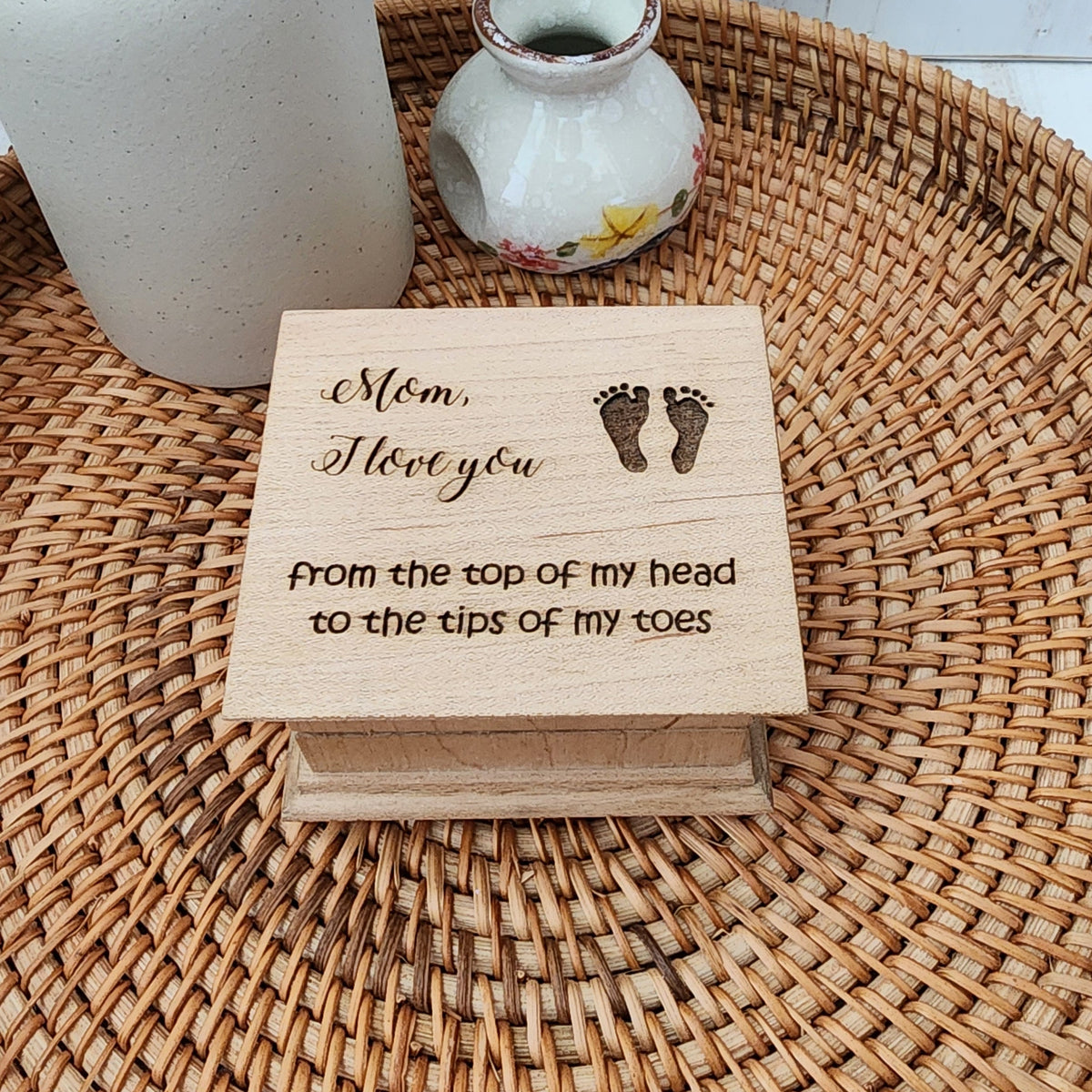 Small wooden music box with Mom I love you from the top of my head to the tips of my toes and footprints on a woven mat with a ceramic vase.