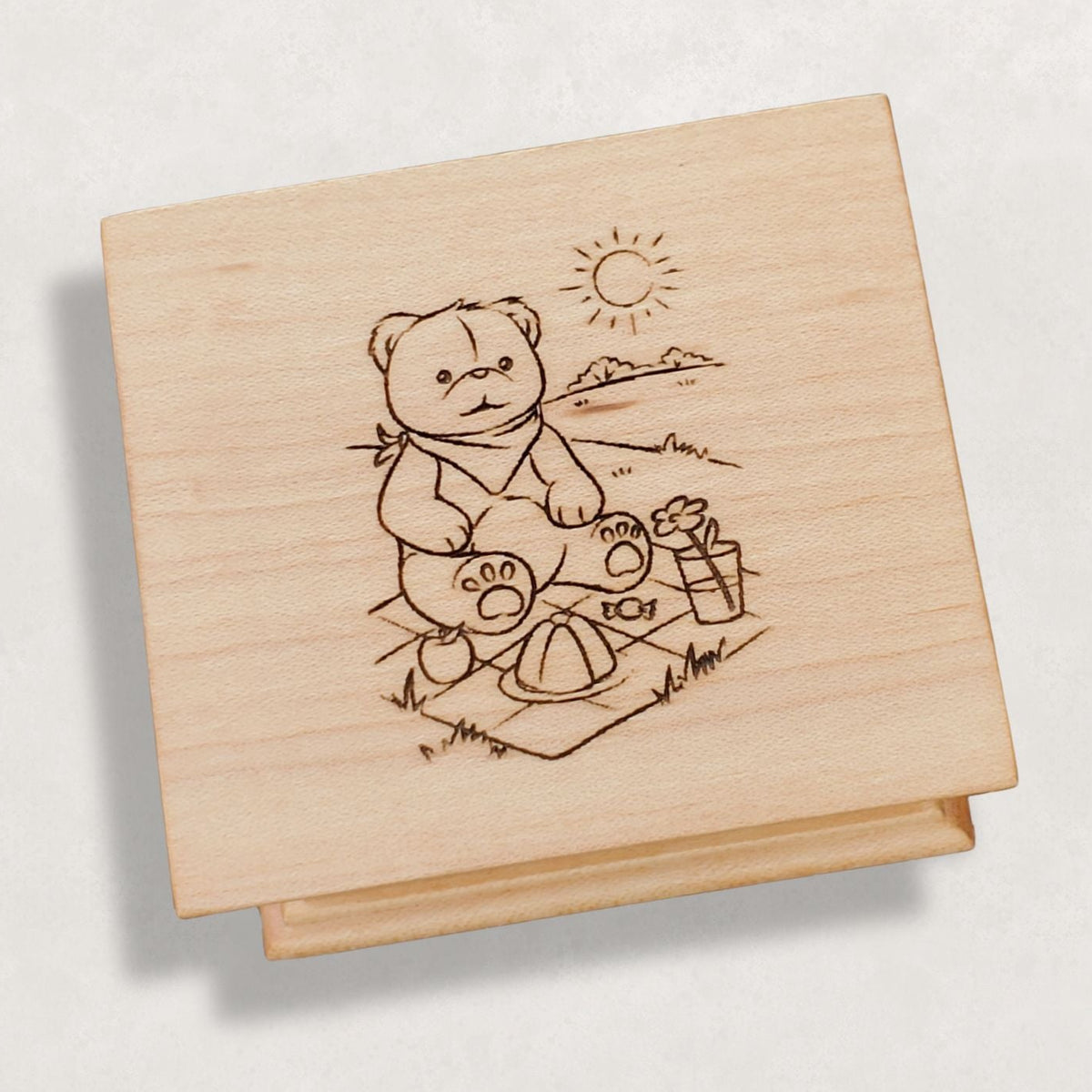Wooden music box with a engraved illustration of a bear having a picnic on a light gray background