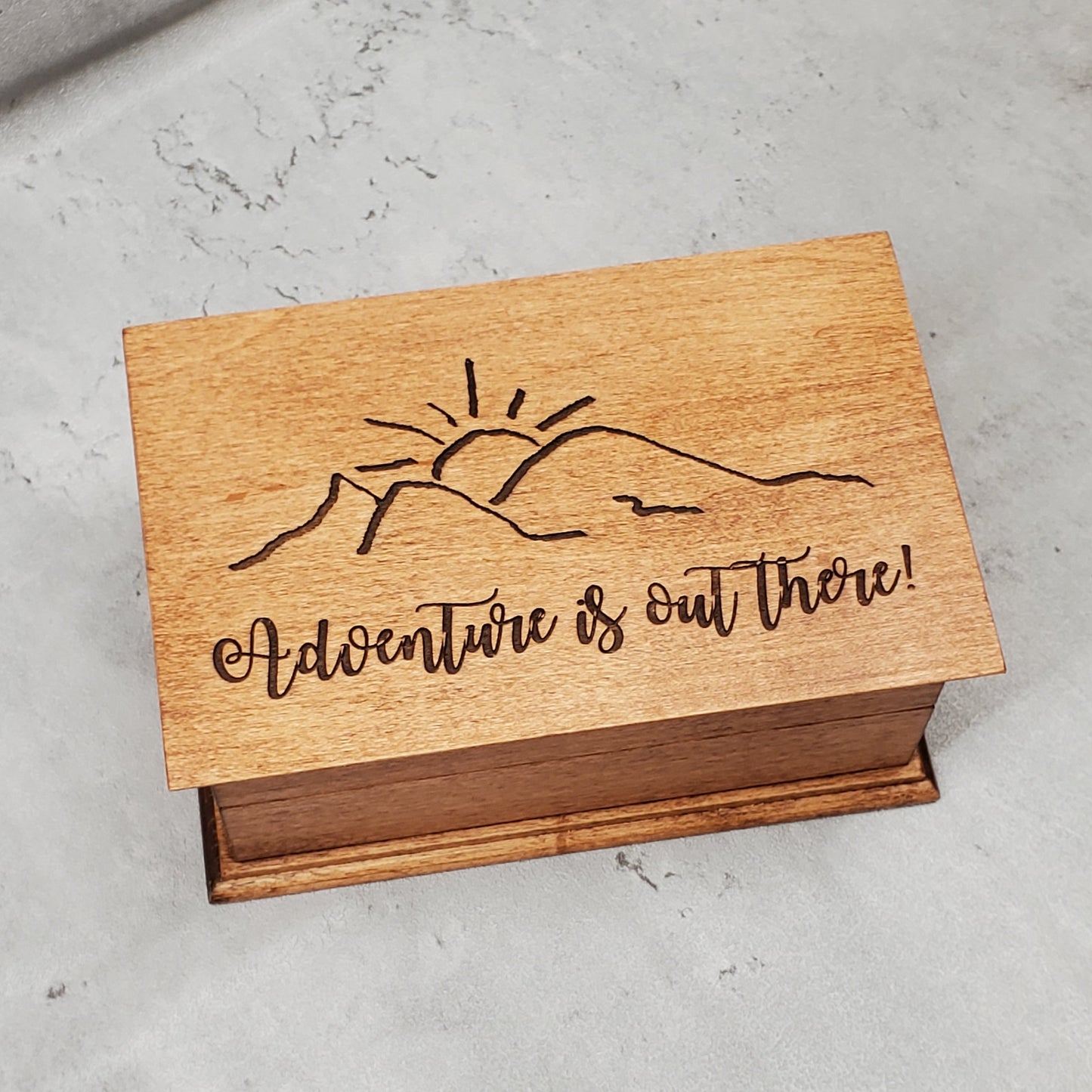 A wooden musical jewelry box with an engraving of a mountain and the text 'Adventure is out there!' on the lid.