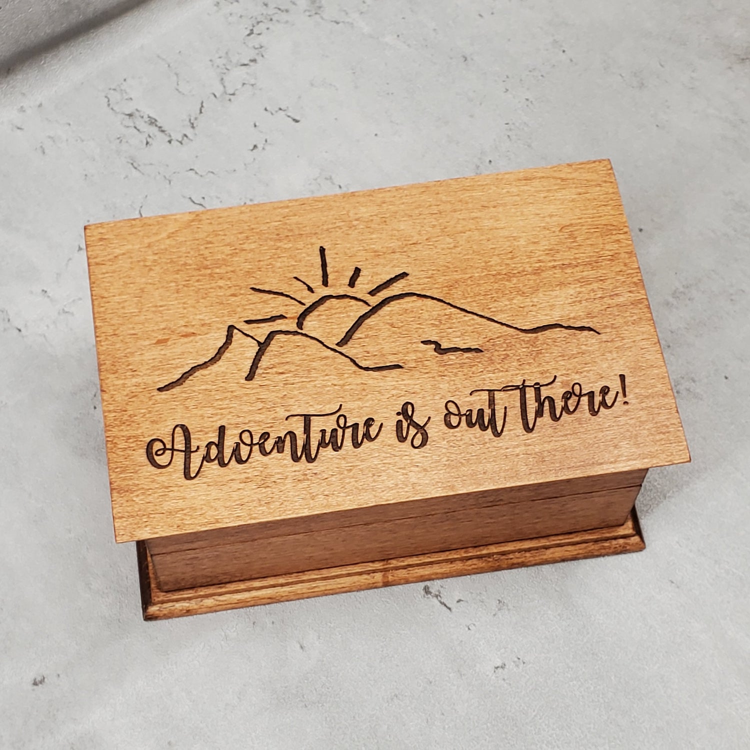 A wooden musical jewelry box with an engraving of a mountain and the text 'Adventure is out there!' on the lid.