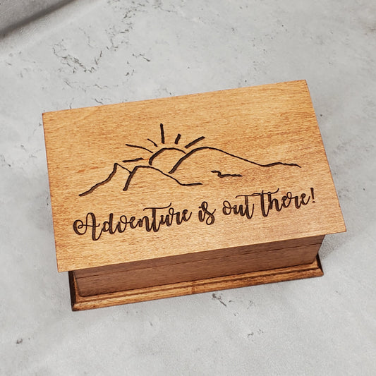 A wooden musical jewelry box with an engraving of a mountain and the text 'Adventure is out there!' on the lid.