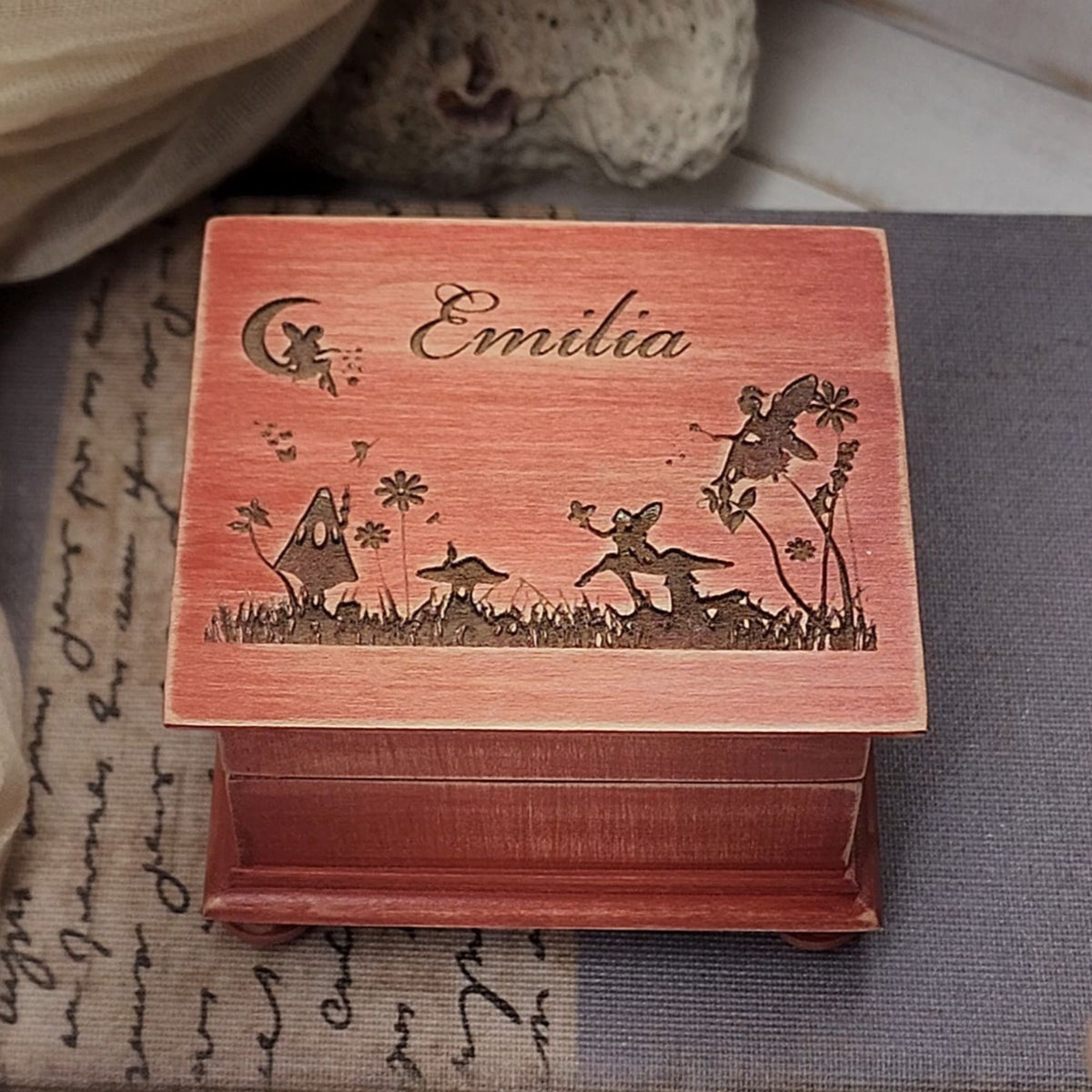 A personalized wooden music box with a reddish-pink lid engraved with the name 'Emilia' and fairy-themed illustrations, shown on a patterned fabric surface.
