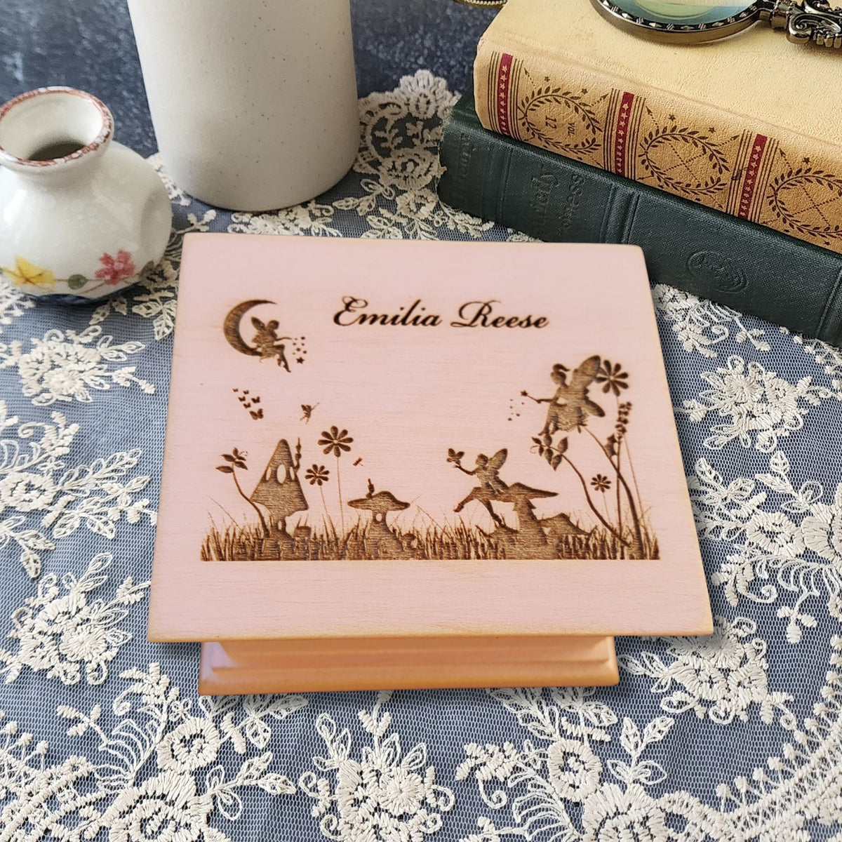 A personalized wooden music box  engraved with fairy and woodland illustrations and the name 'Emilia Reese,' displayed on a lace-covered surface with books and a small vase in the background.