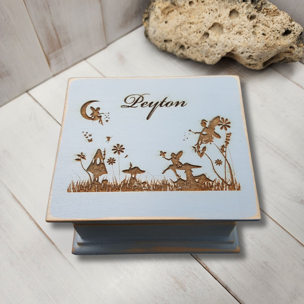 A square wooden music box with a light blue top lid engraved with fairy illustrations, mushrooms, a crescent moon, and the name 'Peyton' in script. The box sits on a whitewashed wooden surface with a natural stone prop in the background.