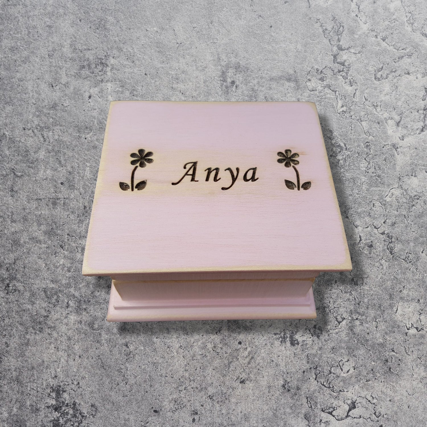 Pink wooden music box with 'Anya' and floral designs on a gray concrete surface