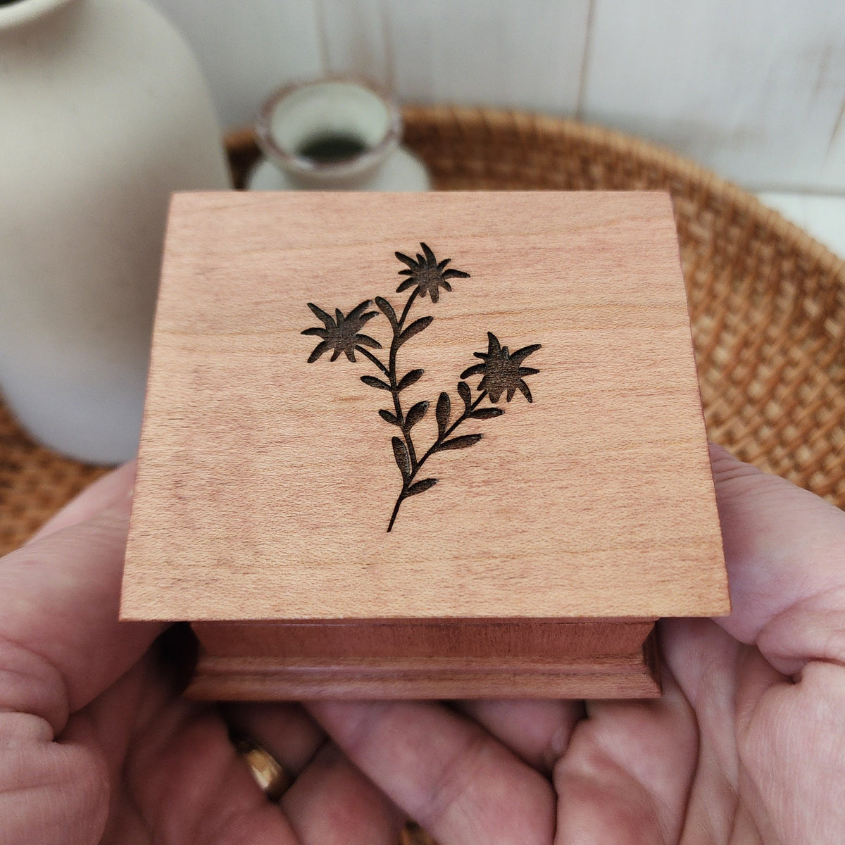 Wooden music box with laser engraved floral design held by a hand, with a blurred background of a white vase and woven basket.
