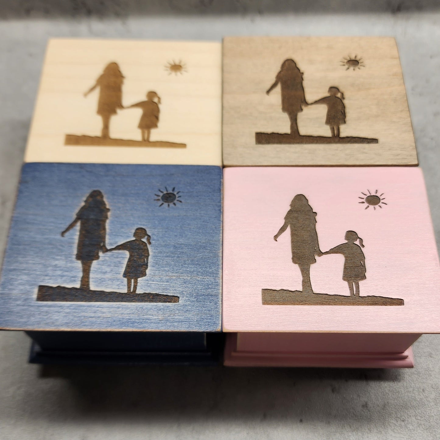Four wooden music boxes in different colors with engraved silhouettes of a woman and a girl walking holding hands on a concrete surface
