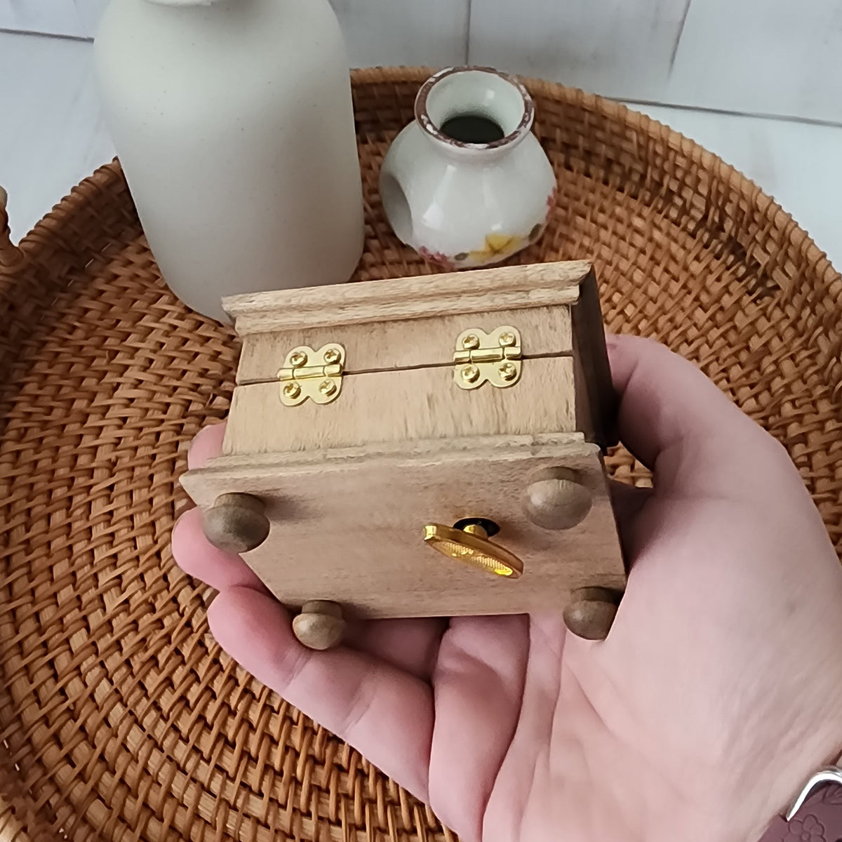 Hand holding a small wooden music box showing the bottom and back side of it with golden wind up key on the bottom and hinges.