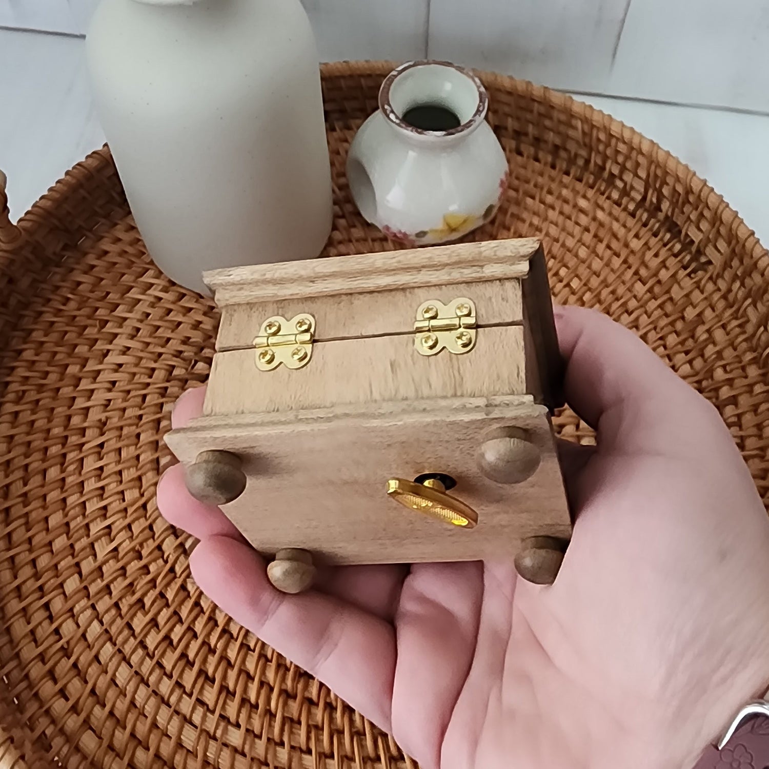 Hand holding a small wooden music box showing the bottom and back side of it with golden wind up key on the bottom and hinges.