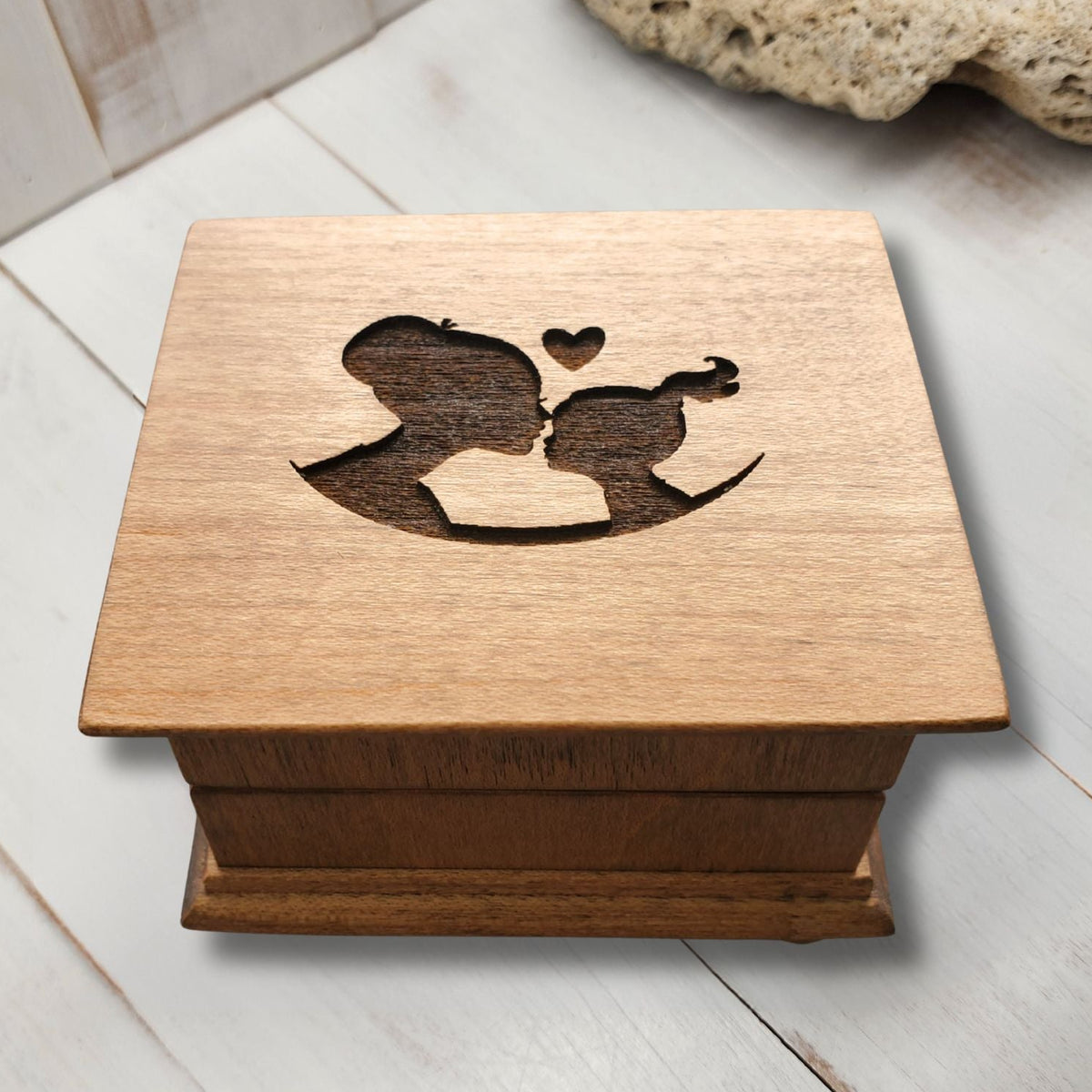 Wooden music box with a laser engraved design of a mom and daughter with a heart above them on a light wooden surface.