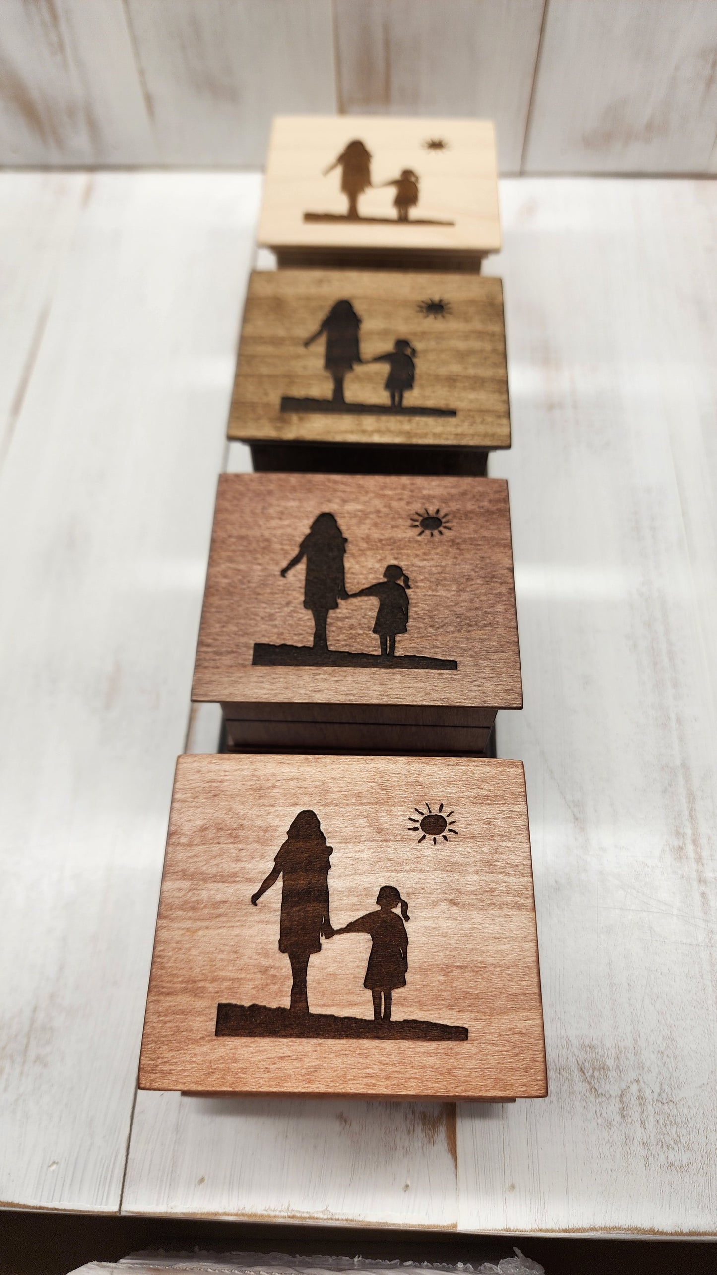 Four wooden music boxes in different colors with engraved silhouettes of a woman and a girl walking holding hands on a concrete surface