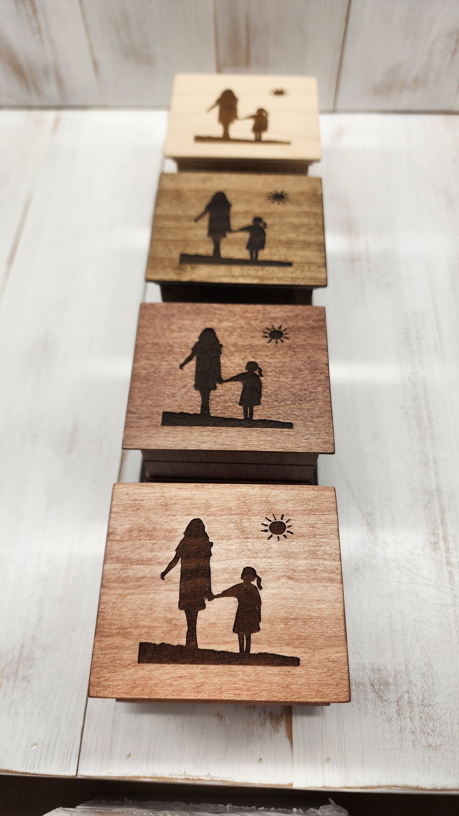 Four wooden music boxes in different colors with engraved silhouettes of a woman and a girl walking holding hands on a concrete surface