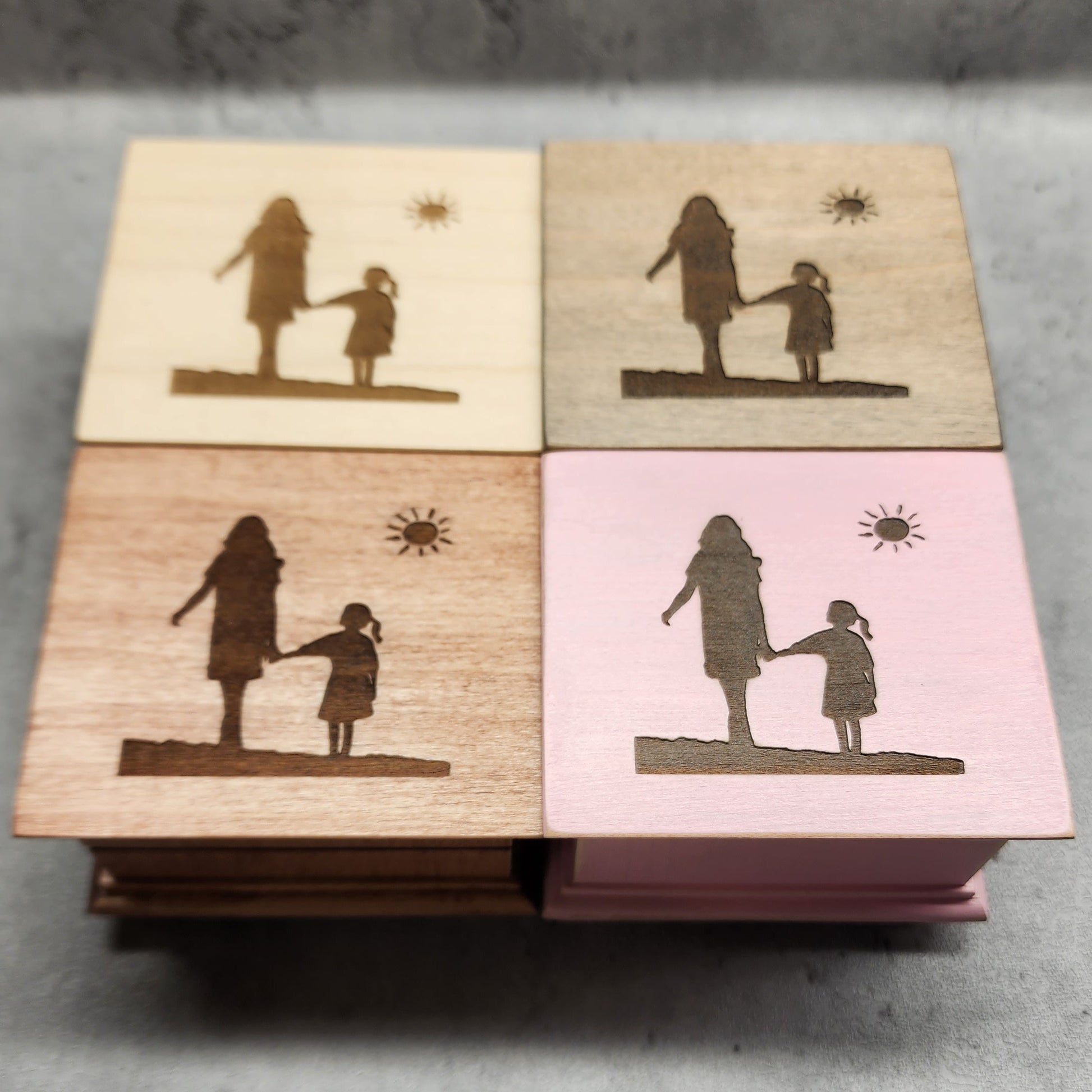 Four wooden music boxes in different colors with engraved silhouettes of a woman and a girl walking holding hands on a concrete surface