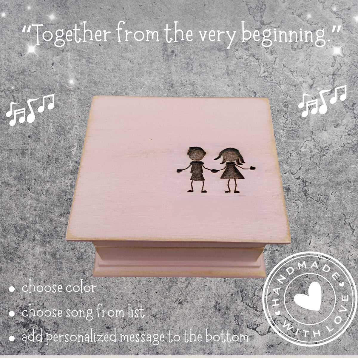 Pink wooden music box with engraved boy and girl silhouette holding hands with a text above the b ox 'Together from the very beginning' on a textured gray background.