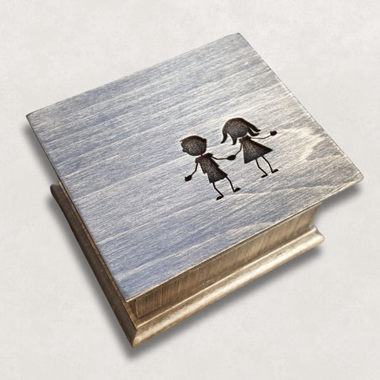 Wooden music box with a silhouette image of a boy and girl holding hands illustration engraved on the top on a light gray background