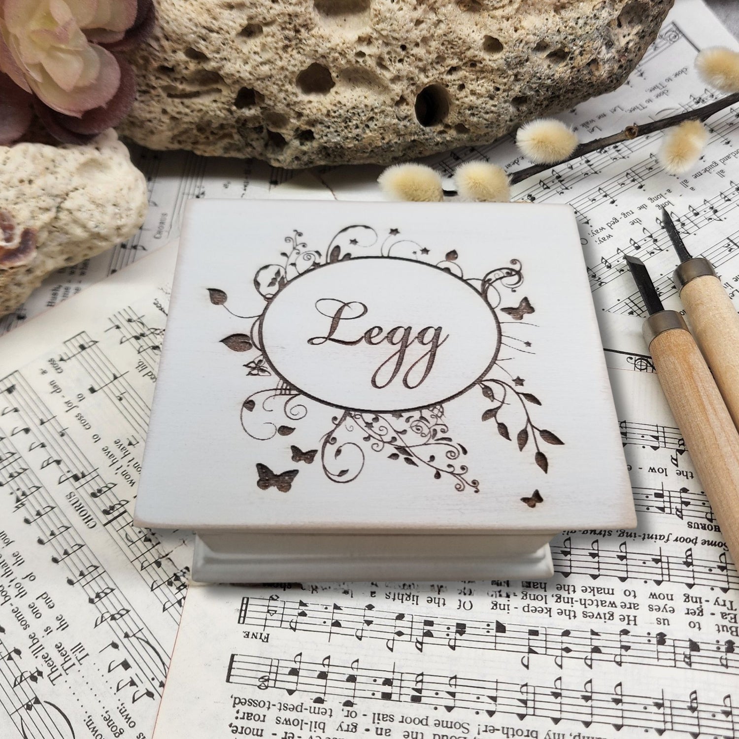 Decorative music box with name 'Legg' engraved on top along with a floral design on sheet music with a rock and flowers in the background