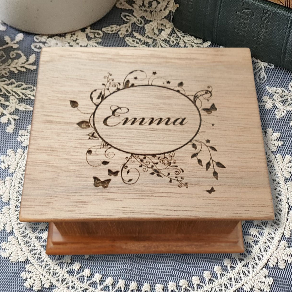 Wooden music box engraved with 'Emma' in a floral oval shaped frame with butterflies on a decorative tablecloth