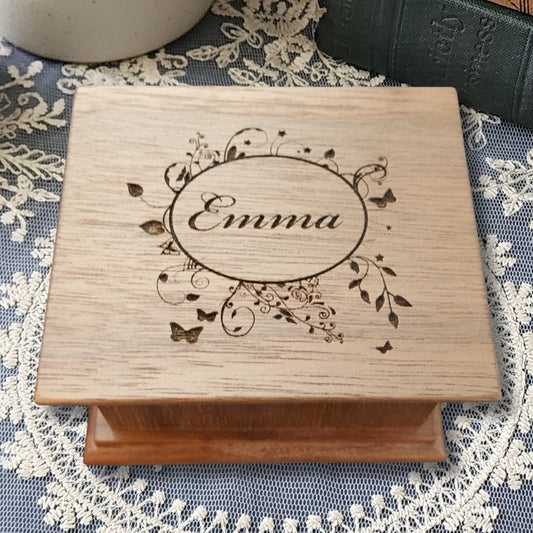 Wooden music box engraved with 'Emma' in a floral oval shaped frame with butterflies on a decorative tablecloth