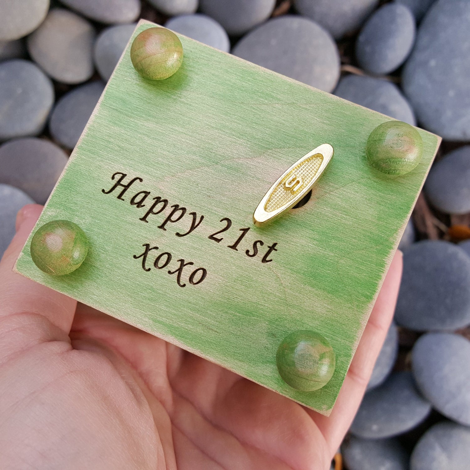 Green wooden card with 'Happy 21st xoxo' text engraved on the bottom side of box, also showing the wind up key and the 4 button feet, a music box is being held over pebbles